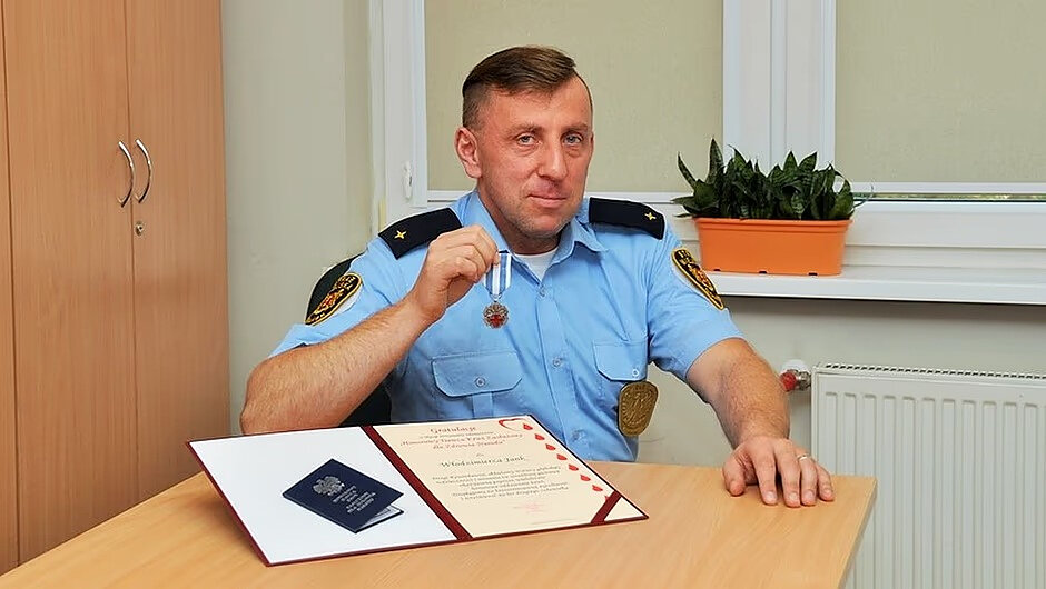 Na zdjęciu widoczna jest osoba siedząca przy biurku w pomieszczeniu przypominającym biuro lub gabinet służbowy. Osoba ma na sobie jasnoniebieską koszulę mundurową z naramiennikami oraz oznaczeniami na rękawach i przypiętą odznaką na piersi. W jednej dłoni osoba trzyma medal na wstążce, prezentując go w kierunku aparatu.
Na biurku przed osobą leży otwarta teczka z dokumentem oraz dwoma ciemnymi książeczkami (prawdopodobnie dokumentami urzędowymi). Dokument zawiera widoczny nagłówek i ozdobne elementy, choć szczegóły tekstu nie są w pełni czytelne. Obok znajduje się także ręka oparta o blat.
W tle znajduje się jasna ściana, szafka z drzwiami po lewej stronie oraz okno z roletą. Na parapecie stoi doniczka z zieloną rośliną. Kolorystyka wnętrza jest stonowana: dominują jasne odcienie beżu, bieli i drewna, co sprawia, że scena ma uporządkowany i formalny charakter.