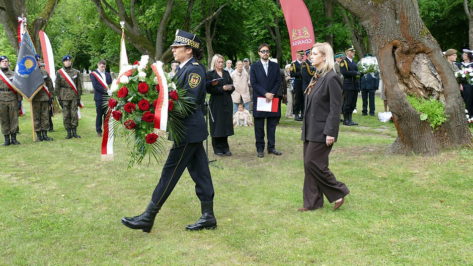 Strażniczka miejska niesie wieniec, za nią idzie Emilia Lodzińska, zastępczyni prezydenta Gdańska. W tle poczty sztandarowe.