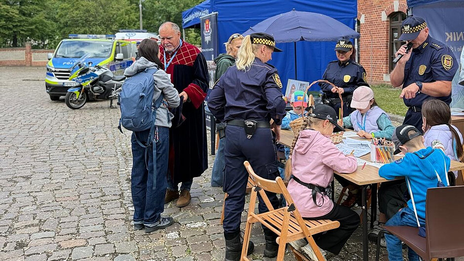 Na zdjęciu przedstawiono scenę plenerowego wydarzenia edukacyjnego, odbywającego się na brukowanym dziedzińcu w pobliżu ceglanego budynku.
? Główna scena
W centralnej części znajduje się drewniany stół, przy którym siedzi kilkoro dzieci. Dzieci wykonują prace plastyczne – kolorują i rysują na kartkach. Na stole ustawiono pojemnik z kolorowymi kredkami, a także koszyk z materiałami.
Obok dzieci stoją funkcjonariusze straży miejskiej ubrani w granatowe mundury z charakterystycznymi czapkami w żółto-czarną kratę. Jedna osoba stoi tyłem do aparatu, druga znajduje się po drugiej stronie stołu i trzyma dokumenty, nadzorując lub pomagając przy zajęciach.
Po prawej stronie kadru widoczna jest osoba w mundurze, która trzyma mikrofon i przemawia, co sugeruje prowadzenie programu wydarzenia lub zapowiedzi dla uczestników.
? Lewa część zdjęcia
Po lewej stronie widoczna jest scena rozmowy: osoba ubrana w bordową togę z ozdobnym wzorem rozmawia z młodą osobą z plecakiem. Obie stoją obok siebie, a ich postawa wskazuje na bezpośrednią interakcję.
W tle znajdują się pojazdy policyjne – samochód oraz motocykl z widocznymi oznaczeniami „Policja”. Pojazdy stoją na kostce brukowej, co dodatkowo podkreśla charakter wydarzenia jako pikniku służb lub dnia otwartego.
? Tło i otoczenie
Za stołem ustawiony jest niebieski namiot (stoisko), przy którym gromadzą się uczestnicy. Wokół widać elementy zieleni – drzewa oraz fragmenty trawnika. Po prawej stronie kadru znajduje się ceglana ściana budynku.
? Podsumowanie
Zdjęcie przedstawia integracyjne wydarzenie z udziałem służb miejskich i mieszkańców. Szczególny nacisk położono na aktywności edukacyjne dla dzieci, prezentację pracy służb oraz bezpośredni kontakt z uczestnikami wydarzenia.
