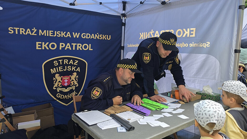 Na trzecim zdjęciu pokazane jest stanowisko informacyjno-edukacyjne Straży Miejskiej w Gdańsku. W tle widoczny jest duży baner z napisem „Straż Miejska w Gdańsku – EKO Patrol” oraz herb miasta. Przy stole siedzi dwóch funkcjonariuszy w charakterystycznych mundurach, którzy prezentują dzieciom przygotowane materiały edukacyjne, takie jak kartki, plakietki czy kolorowe paski papieru. Na stole znajdują się także dokumenty, artykuły biurowe oraz pudełko z materiałami. Dwoje dzieci stoi po drugiej stronie stołu i przygląda się prezentacji. Cała scena ma charakter edukacyjny i promuje działania ekologiczne oraz bezpieczeństwo.