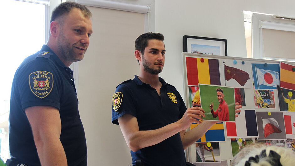 Na zdjęciu widoczna jest scena wewnątrz jasnego pomieszczenia z białymi ścianami i oknem zasłoniętym roletą. W pierwszym planie, po lewej stronie, stoi jedna osoba w granatowej koszulce z krótkim rękawem, na której znajduje się wyraźna naszywka z napisem „STRAŻ MIEJSKA GDAŃSK”. Obok, w centralnej części kadru, znajduje się druga osoba ubrana podobnie, również z oznaczeniem tej samej instytucji na rękawie. Druga osoba trzyma przed sobą planszę lub tablicę prezentacyjną, na której przypięte są różne zdjęcia oraz ilustracje przedstawiające flagi państw i sceny sportowe. Wśród nich widoczna jest flaga Niemiec, Japonii i Francji, a także inne zdjęcia, w tym fotografia sportowca na boisku. Elementy na tablicy są rozmieszczone w sposób uporządkowany i przymocowane kolorowymi pinezkami. W dolnej części zdjęcia widać fragmenty głów osób siedzących, co sugeruje obecność publiczności słuchającej prezentacji. W tle znajdują się także elementy wyposażenia pomieszczenia, takie jak ramka z obrazem na ścianie oraz drzwi po prawej stronie. Oświetlenie jest naturalne i jasne, co pozwala wyraźnie dostrzec szczegóły ubioru, tablicy oraz otoczenia. Cała scena przedstawia sytuację edukacyjną lub informacyjną, w której osoby stojące prowadzą prezentację dla zgromadzonej grupy.