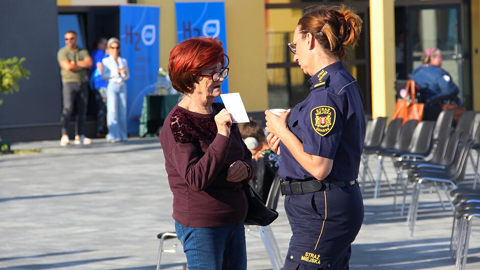 Na zdjęciu przedstawiona jest scena z wydarzenia plenerowego odbywającego się na otwartym, wybrukowanym placu w słoneczny dzień. Na pierwszym planie widoczne są dwie osoby prowadzące rozmowę — jedna ubrana w granatowy mundur z naszywką „STRAŻ MIEJSKA GDAŃSK” na rękawie, druga w codzienny strój składający się z ciemnej bluzki i spodni. Osoba w mundurze trzyma w dłoni mały papierowy kubek, natomiast druga osoba trzyma niewielką kartkę i zwrócona jest w stronę rozmówcy. W tle widoczne są rzędy ustawionych krzeseł, a także kilka osób stojących i rozmawiających w pobliżu stanowisk informacyjnych. Z lewej strony znajdują się stoliki z materiałami edukacyjnymi oraz pionowe banery z widocznymi napisami, w tym „H₂O”, co wskazuje na tematykę związaną z wodą lub ochroną środowiska. Za uczestnikami widać również fragmenty budynku o jasnej elewacji oraz elementy infrastruktury wydarzenia. Całość oświetlona jest intensywnym światłem słonecznym, które tworzy wyraźne cienie i podkreśla szczegóły ubioru, przedmiotów oraz otoczenia.