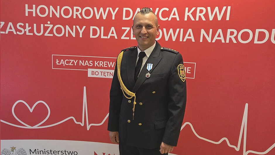 Na tle okolicznościowej ścianki promującej honorowe krwiodawstwo stoi strażak w mundurze galowym, udekorowany odznaką „Honorowy Dawca Krwi – Zasłużony dla Zdrowia Narodu”, przyznaną za wieloletnie, bezinteresowne oddawanie krwi.