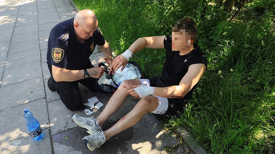 Na zdjęciu przedstawiono sytuację udzielania pomocy na chodniku biegnącym wzdłuż drogi, w otoczeniu zieleni. W centralnej części kadru widoczna jest osoba siedząca na krawężniku, ubrana w czarną koszulkę bez rękawów, jasne spodenki i sportowe buty, z widocznymi otarciami i zadrapaniami na nogach oraz zabandażowanymi przedramionami i kolanem. Obok tej osoby klęczy funkcjonariusz Straży Miejskiej w granatowym mundurze z naszywkami i emblematem, który opatruje ranę, korzystając z elementów znajdujących się w torbie medycznej. Na ziemi wokół znajdują się zużyte opatrunki oraz plastikowa butelka z wodą. Miejsce zdarzenia oświetlone jest silnym światłem słonecznym, przez co widoczne są wyraźne cienie drzew i osób, a tło stanowi gęsta, zielona roślinność rosnąca wzdłuż chodnika.
