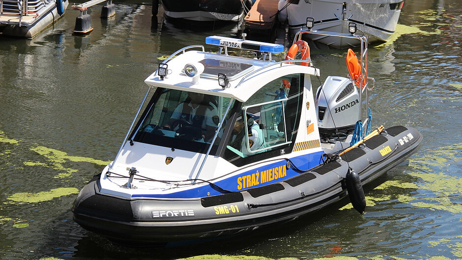 Copilot said: Na zdjęciu widoczna jest nowoczesna łódź patrolowa unosząca się na spokojnej wodzie w porcie lub marinie. Jednostka ma biało‑czarną konstrukcję z niebieskimi i żółtymi elementami oraz wyraźnym napisem „STRAŻ MIEJSKA” umieszczonym na burcie. W przedniej części łodzi znajduje się zamknięta kabina z dużymi szybami, przez które widać osoby siedzące wewnątrz. Na dachu kabiny zamontowano sygnały świetlne, reflektory oraz dodatkowy sprzęt.
Z tyłu łodzi widoczny jest silnik zaburtowy marki „HONDA”, a także elementy wyposażenia ratunkowego, w tym pomarańczowe koła ratunkowe i metalowa konstrukcja zabezpieczająca. Kadłub łodzi ma pompowane burty, co wskazuje na typ RIB (Rigid Inflatable Boat), przystosowany do działań patrolowych i ratowniczych.
Woda wokół jednostki ma zielonkawy odcień, z widocznymi skupiskami roślinności unoszącej się na powierzchni. W tle znajdują się inne zacumowane łodzie oraz elementy infrastruktury portowej, takie jak pomosty i pływające platformy. Cała scena jest dobrze oświetlona naturalnym światłem dziennym, które podkreśla szczegóły konstrukcji łodzi i odbicia na wodzie.