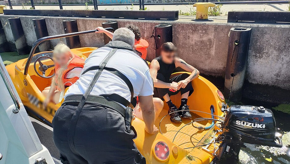 Copilot said: Na zdjęciu przedstawiona jest scena odbywająca się na wodzie, przy betonowym nabrzeżu z widocznymi cylindrycznymi odbojnikami oraz metalową barierką w tle. Na pierwszym planie znajduje się niewielka łódź o intensywnym, żółto‑pomarańczowym kolorze, częściowo zanurzona w wodzie pokrytej fragmentami roślinności. Na rufie zamontowany jest silnik zaburtowy z widocznym napisem „SUZUKI”.
W łodzi przebywa kilka osób – część z nich ma na sobie pomarańczowe kamizelki ratunkowe. Twarze osób zostały celowo rozmyte. Jedna osoba siedzi z przodu przy kierownicy łodzi, inna siedzi z tyłu, trzymając dłonie razem, a jeszcze jedna znajduje się pomiędzy nimi. Obok łodzi, częściowo w kadrze, widoczna jest druga jednostka pływająca.
Najbliżej aparatu znajduje się osoba stojąca na brzegu lub na sąsiedniej łodzi, pochylona nad burtą i wykonująca czynności związane z pomocą lub obsługą łodzi. Osoba ta ma na sobie jasną koszulkę oraz uprząż lub pas zabezpieczający przypięty do ciała. Widoczny jest także fragment sprzętu przypiętego do pasa.
Cała scena jest dobrze oświetlona, co sugeruje słoneczny dzień. Kolory są wyraźne i kontrastowe – szczególnie wyróżnia się jasna łódź oraz pomarańczowe kamizelki ratunkowe na tle szarego betonu i ciemniejszej wody.