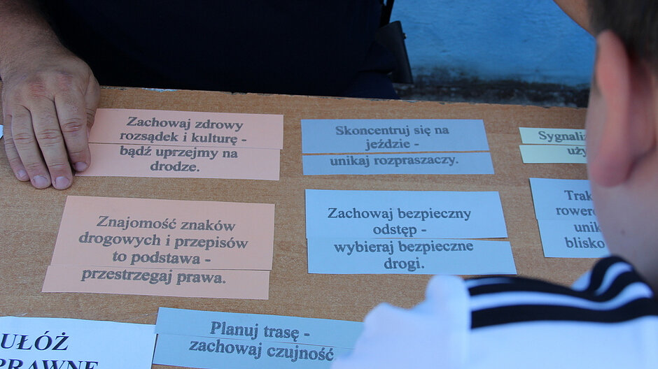 Na zdjęciu widoczny jest fragment drewnianego stołu, nad którym znajduje się osoba po jednej stronie oraz druga osoba po przeciwnej stronie, częściowo widoczna z tyłu głowy i ramienia; na blacie stołu rozmieszczone są prostokątne paski papieru w różnych kolorach (m.in. jasnoróżowe i jasnoniebieskie) z wydrukowanymi zasadami dotyczącymi bezpieczeństwa, takimi jak „Zachowaj zdrowy rozsądek i kulturę – bądź uprzejmy na drodze.”, „Znajomość znaków drogowych i przepisów to podstawa – przestrzegaj prawa.”, „Skoncentruj się na jeździe – unikaj rozpraszaczy.” oraz „Zachowaj bezpieczny odstęp – wybieraj bezpieczne drogi.”; jedna z osób trzyma dłoń przy stole, jakby wskazując lub układając elementy, a całość jest oświetlona naturalnym światłem, które podkreśla kontrast między jasnymi kartkami z tekstem a brązową powierzchnią stołu.