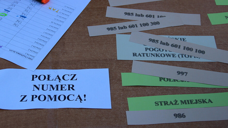 Na zdjęciu widoczny jest fragment drewnianego stołu, na którym rozłożone są paski papieru z wydrukowanymi numerami alarmowymi oraz nazwami służb i instytucji, a także kartka z dużym napisem „POŁĄCZ NUMER Z POMOCĄ!”; obok znajduje się arkusz z tabelą i zapisanymi odręcznie wynikami zaznaczonymi kolorowymi markerami oraz leżący zakreślacz, a jedna dłoń opiera się o kartkę, co sugeruje wykonywanie zadania polegającego na dopasowywaniu numerów telefonów do odpowiednich służb; całość oświetlona jest naturalnym światłem, które podkreśla kontrast między jasnymi kartkami a brązową powierzchnią stołu.