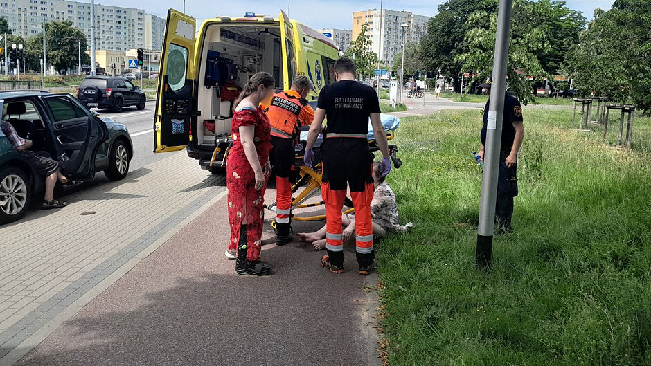 Na poboczu stoi samochód, obok karetka pogotowia z otwartymi drzwiami. Obok karetki, plecami do obiektywu, stoi dwóch ratowników medycznych, obok kobieta, a za latarnią strażnik miejski. Na trawie natomiast siedzi kobieta, której wszyscy udzielają pomocy. 