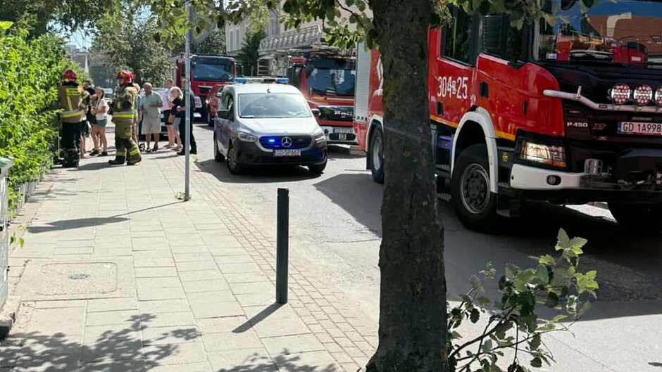 Zaparkowane na ulicy - trzy wozy straży pożarnej oraz jeden radiowóz straży miejskiej. W tle po lewej stronie widoczni także strażacy i kilkoro mieszkańców. 