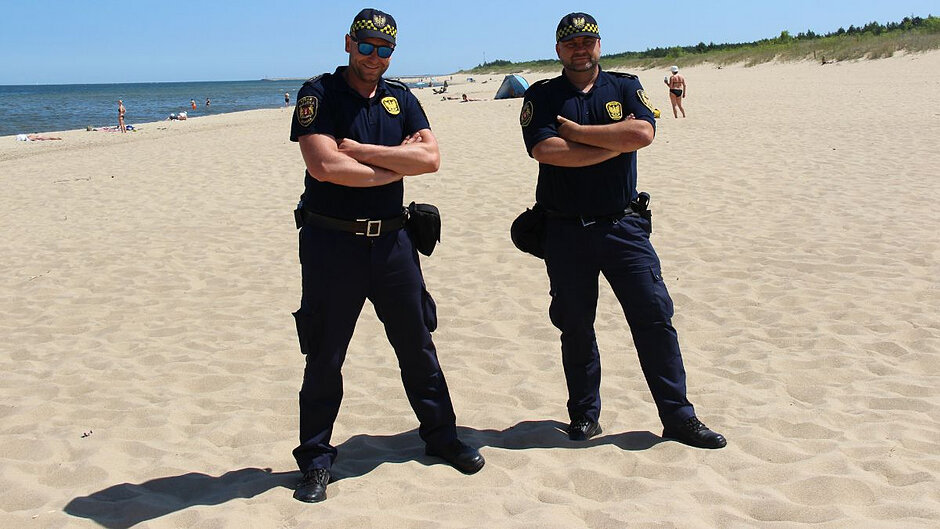 Dwóch strażników miejskich stoi na plaży. Za nimi w tle plażowicze. 