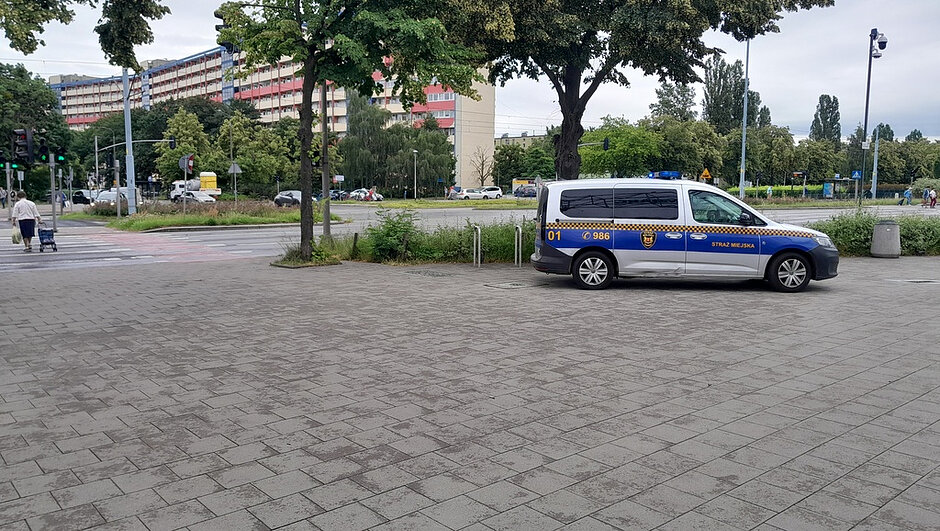 Na zdjęciu widoczny jest oznakowany pojazd Straży Miejskiej zaparkowany na utwardzonym placu wyłożonym szarą kostką brukową, w pobliżu skrzyżowania z przejściem dla pieszych. Na pojeździe włączone są niebieskie sygnały świetlne. W tle znajdują się drzewa, pas zieleni, jezdnia z poruszającymi się pojazdami oraz wielokondygnacyjny budynek mieszkalny. Pogoda jest pochmurna, nawierzchnia wygląda na suchą.