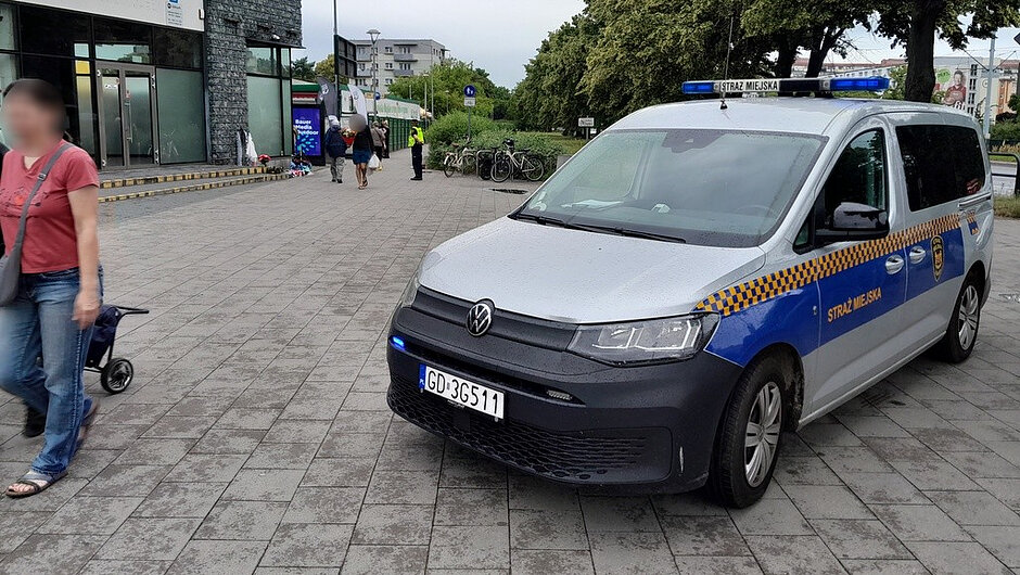 Zdjęcie przedstawia oznakowany pojazd Straży Miejskiej marki Volkswagen zaparkowany na chodniku przed budynkiem usługowym. Na dachu oraz z przodu pojazdu widoczne są włączone niebieskie sygnały świetlne. W pobliżu znajdują się piesi, stojaki rowerowe z rowerami oraz wejście do budynku. Na pojeździe widoczny jest napis „STRAŻ MIEJSKA” oraz numer rejestracyjny zaczynający się od „GD”. Nawierzchnia wykonana jest z kostki brukowej.