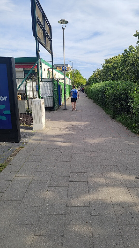 Na zdjęciu widoczny jest szeroki chodnik przebiegający pomiędzy budynkiem handlowo-usługowym a pasem wysokiej zieleni. Przy budynku znajduje się słup reklamowy oraz ogrodzenie. Chodnikiem poruszają się piesi, a w dalszej części widoczne są kolejne osoby. Pośrodku ciągu pieszego znajduje się latarnia uliczna oraz znak drogowy nakazujący ruch dla rowerów. Zdjęcie wykonano w ciągu dnia przy dobrej widoczności i słonecznej pogodzie.