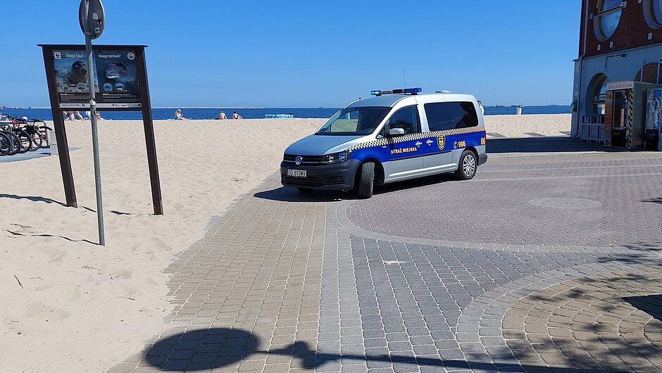 Na zdjęciu przedstawiono nadmorską promenadę przy plaży. W centralnej części kadru znajduje się oznakowany pojazd Straży Miejskiej, zaparkowany na utwardzonej nawierzchni przy wejściu na plażę. Po lewej stronie widoczne są tablice informacyjne oraz stojaki z rowerami. W tle znajduje się piaszczysta plaża, morze i osoby wypoczywające nad wodą. Po prawej stronie widoczny jest fragment budynku położonego przy promenadzie. Zdjęcie wykonano w słoneczny dzień przy dobrej widoczności, co pozwala na wyraźne ukazanie infrastruktury plażowej oraz działań patrolowych służb miejskich w rejonie wypoczynkowym.