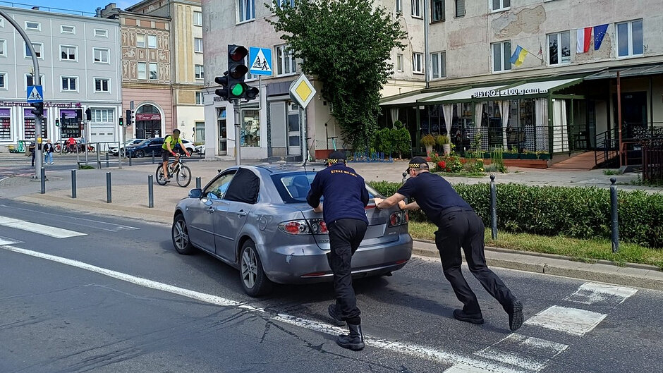 Na zdjęciu widoczna jest miejska ulica w centrum miasta. Na pierwszym planie znajdują się dwie osoby w granatowych mundurach z napisem „STRAŻ MIEJSKA”, które pchają srebrny samochód osobowy stojący na jezdni. Pojazd znajduje się w pobliżu przejścia dla pieszych i skrzyżowania z sygnalizacją świetlną. Światło dla pojazdów jest zielone.
Samochód ma zasłonięte ciemnym materiałem boczne okno po lewej stronie oraz widoczne uszkodzenia i otarcia lakieru na tylnym lewym błotniku. Funkcjonariusze pchają auto od tyłu, prawdopodobnie usuwając je z pasa ruchu.
W tle widać zabudowę miejską z kilkupiętrowymi kamienicami oraz lokal gastronomiczny z ogródkiem. Na elewacji budynku widoczne są flagi, w tym flaga Polski i Unii Europejskiej. Po lewej stronie zdjęcia znajduje się rowerzysta przejeżdżający przez skrzyżowanie. Wzdłuż chodnika ustawiono słupki zabezpieczające oraz znaki drogowe, w tym znak przejścia dla pieszych i znak drogi z pierwszeństwem przejazdu.
Zdjęcie wykonano w ciągu dnia przy dobrej widoczności. Cała scena przedstawia sytuację usuwania unieruchomionego pojazdu z ruchliwej ulicy przez straż miejską.
