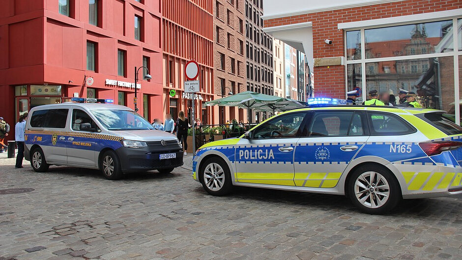 Na zdjęciu widoczne są oznakowane pojazdy Policji i Straży Miejskiej podczas wspólnych działań prowadzonych w centrum Gdańska. W pobliżu radiowozów funkcjonariusze realizują czynności służbowe w przestrzeni publicznej.