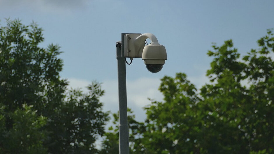 Na zdjęciu przedstawiono kamerę monitoringu wizyjnego zamontowaną na metalowym słupie. Kamera typu obrotowego znajduje się w centralnej części kadru i jest skierowana w dół. W tle widoczne są zielone korony drzew oraz jasne, lekko zachmurzone niebo. Fotografia została wykonana w plenerze, przy naturalnym oświetleniu dziennym. Dominującymi kolorami są odcienie zieleni, szarości i błękitu. Zdjęcie przedstawia element infrastruktury monitoringu zainstalowany na terenie zewnętrznym otoczonym roślinnością.