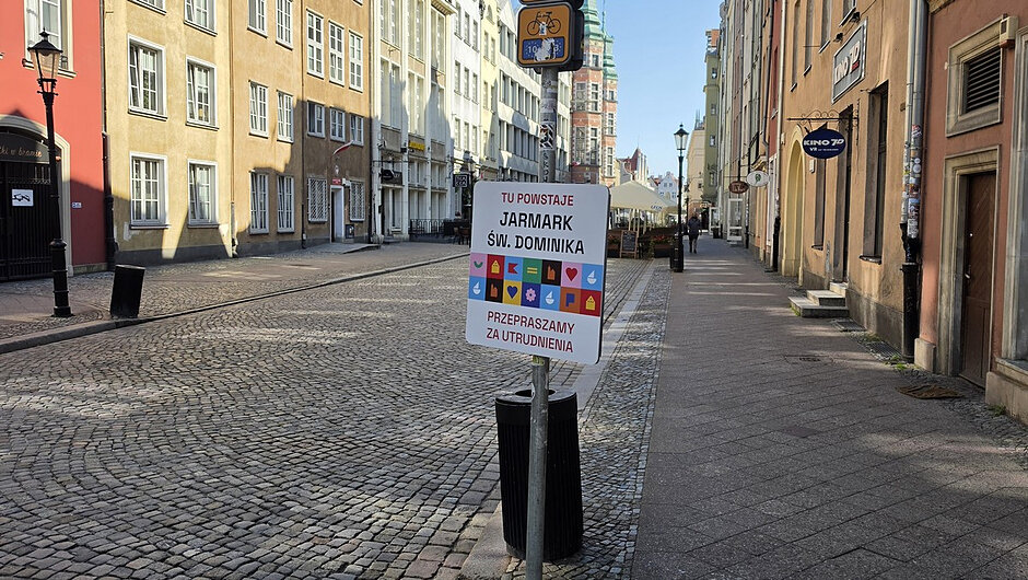 Na drugim zdjęciu przedstawiono ulicę wyłączoną z ruchu na potrzeby organizacji wydarzenia. Na środku znajduje się tablica informacyjna z napisem:
TU POWSTAJE JARMARK ŚW. DOMINIKA. PRZEPRASZAMY ZA UTRUDNIENIA.
Ulica jest wolna od ruchu samochodowego, co wskazuje na prowadzone prace przygotowawcze lub montaż infrastruktury jarmarkowej.
Na trzecim zdjęciu widoczny jest pojazd Straży Miejskiej oraz funkcjonariusz kierujący ruchem przy użyciu tarczy do zatrzymywania pojazdów. Sytuacja wskazuje na prowadzenie zabezpieczenia organizacji ruchu i kontrolę dostępu do strefy objętej czasowymi ograniczeniami.