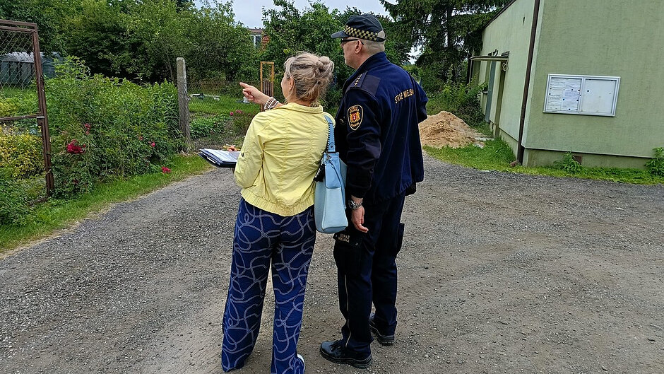 Na zdjęciu przedstawiono teren ogrodów działkowych. Na pierwszym planie, na żwirowej drodze, stoją dwie osoby prowadzące rozmowę i obserwujące teren znajdujący się po lewej stronie kadru. Jedna z osób wskazuje ręką w kierunku działek. Druga osoba ubrana jest w granatowy strój z wyraźnym napisem „STRAŻ MIEJSKA” na plecach.
Po prawej stronie znajduje się budynek o jasnozielonej elewacji, na którego ścianie zamontowana jest widoczna na pierwszym zdjęciu gablotka ogłoszeniowa. Obok budynku widać pryzmę piasku.
W tle znajdują się liczne drzewa, krzewy oraz ogrodzone działki z nasadzeniami i kwiatami. Niebo jest zachmurzone, a światło rozproszone, co wskazuje na pochmurną pogodę. Zdjęcie dokumentuje wizytę lub czynności kontrolne prowadzone na terenie rodzinnych ogrodów działkowych.