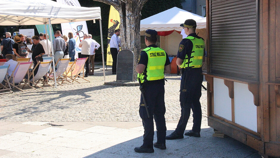 Na zdjęciu widocznych jest dwóch funkcjonariuszy Straży Miejskiej pełniących służbę podczas wydarzenia plenerowego. Funkcjonariusze obserwują teren imprezy, na którym znajdują się uczestnicy, namioty wystawowe, stoiska oraz leżaki przeznaczone do wypoczynku. Fotografia dokumentuje działania związane z zapewnieniem bezpieczeństwa i porządku publicznego podczas wydarzenia.