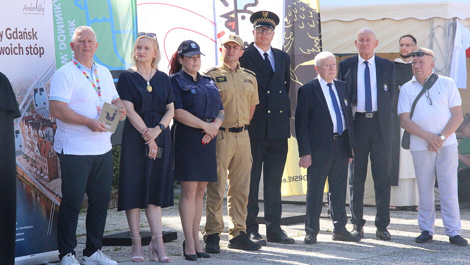 Na fotografii widoczna jest grupa uczestników uroczystości towarzyszącej wydarzeniom związanym z Jarmarkiem św. Dominika. Osoby ustawione są do wspólnego zdjęcia na tle banerów promocyjnych i flag wydarzenia. Wśród zgromadzonych znajdują się przedstawiciele różnych służb, instytucji oraz organizatorów. Zdjęcie dokumentuje oficjalną część spotkania i współpracę podmiotów zaangażowanych w organizację przedsięwzięcia.