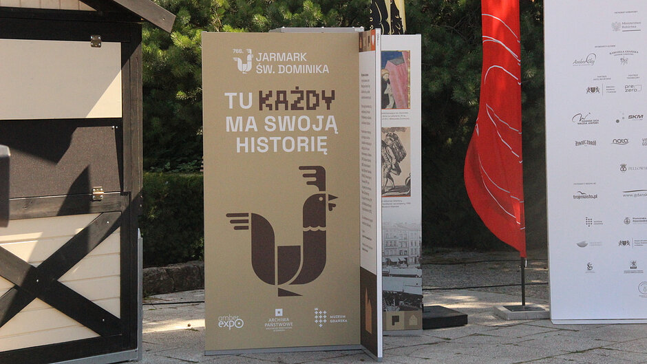 Fotografia przedstawia planszę wystawową promującą 766. Jarmark św. Dominika. Na ekspozycji widoczny jest napis „TU KAŻDY MA SWOJĄ HISTORIĘ” oraz materiały związane z historią i tradycją wydarzenia. Plansza stanowi element wystawy plenerowej przygotowanej dla uczestników i odwiedzających.