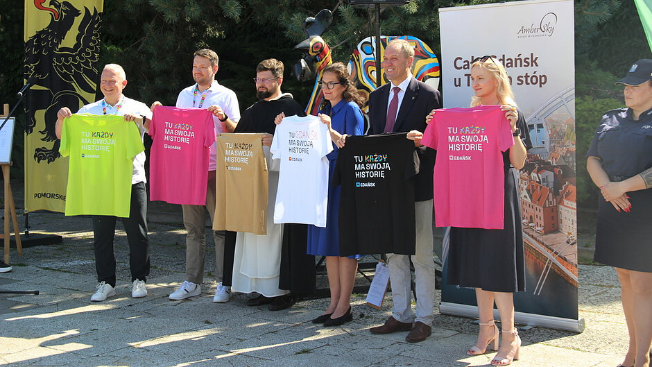 Na zdjęciu uczestnicy wydarzenia prezentują koszulki promocyjne z hasłem „TU KAŻDY MA SWOJĄ HISTORIĘ”. Fotografia została wykonana podczas oficjalnej części wydarzenia i dokumentuje działania promocyjne związane z Jarmarkiem św. Dominika. W tle widoczne są banery informacyjne oraz elementy identyfikacji wizualnej miasta Gdańska i organizatorów przedsięwzięcia.