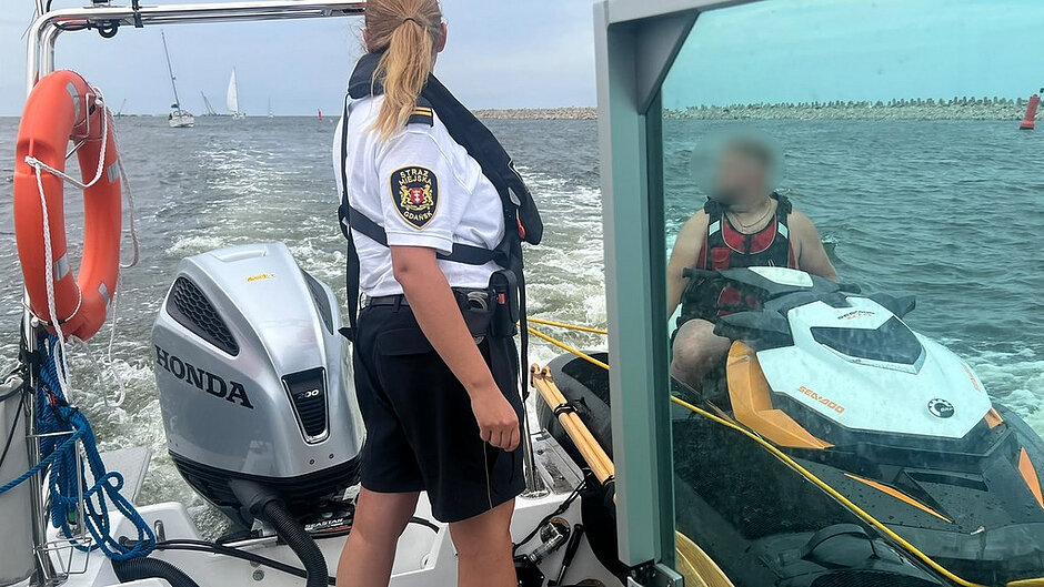 Na zdjęciu widoczna jest łódź patrolowa Straży Miejskiej płynąca po akwenie wodnym. Na pokładzie, przy rufie jednostki, znajduje się funkcjonariusz Straży Miejskiej ubrany w kamizelkę ratunkową i umundurowanie służbowe z emblematem „Straż Miejska Gdańsk”. Obok widoczny jest silnik zaburtowy marki Honda oraz koło ratunkowe zamocowane do konstrukcji łodzi. Po prawej stronie zdjęcia znajduje się skuter wodny marki Sea-Doo, przy którym przebywa osoba w kamizelce asekuracyjnej. Pomiędzy łodzią patrolową a skuterem widoczna jest lina zabezpieczająca. W tle znajdują się falochron, oznakowanie nawigacyjne oraz żaglówki poruszające się po wodzie.
