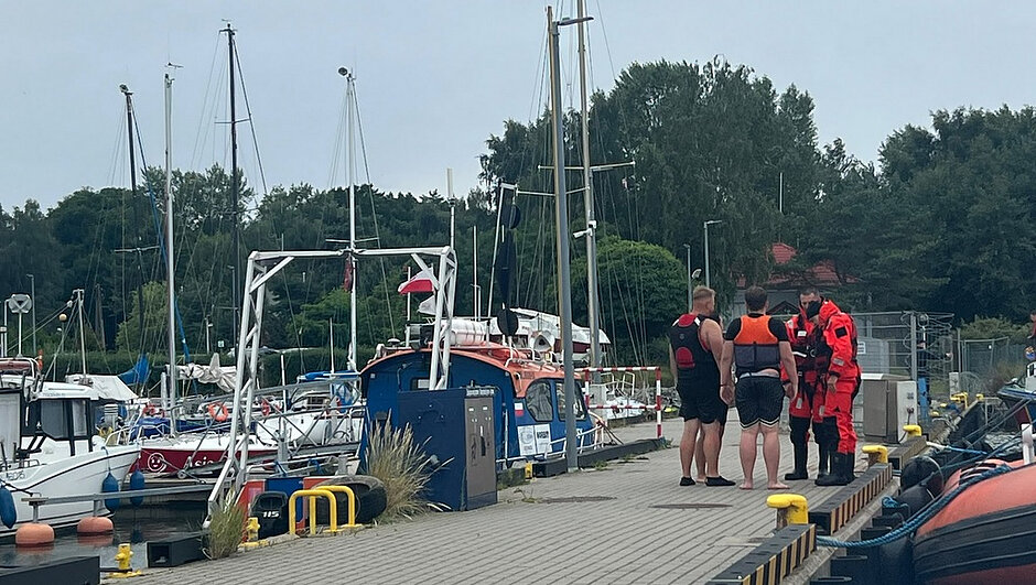 Na zdjęciu przedstawiono pomost w marinie, przy którym prowadzone są czynności po dopłynięciu do nabrzeża. Na pomoście stoją dwie osoby ubrane w kamizelki asekuracyjne oraz dwie osoby w specjalistycznych czerwono-czarnych kombinezonach ratowniczych. W tle widoczne są zacumowane jachty żaglowe, łodzie motorowe oraz infrastruktura portowa. Po prawej stronie kadru znajduje się fragment łodzi patrolowej z pontonową burtą. Na zdjęciu widoczne są również urządzenia techniczne nabrzeża, polery cumownicze oraz elementy zabezpieczające pomost. Całość przedstawia działania służb prowadzone na terenie mariny po zakończeniu interwencji na wodzie.