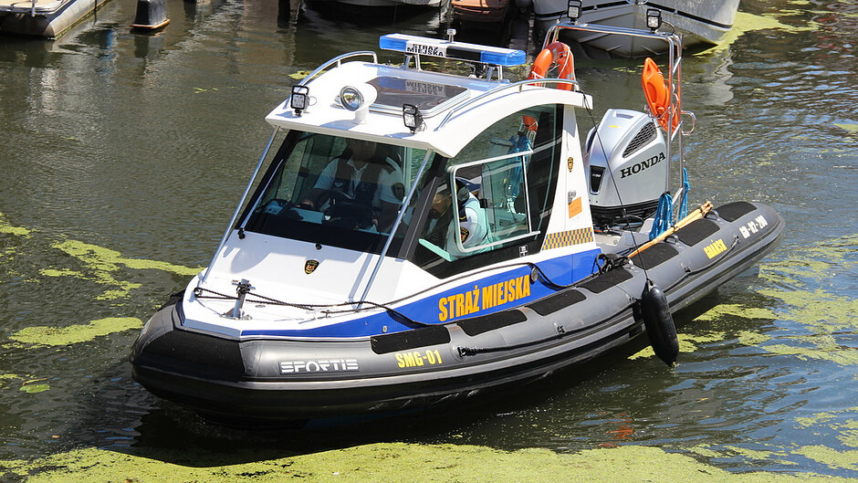 Na zdjęciu przedstawiono patrolową łódź Straży Miejskiej typu RIB z zamkniętą kabiną, znajdującą się na wodach portowych lub w marinie. Jednostka ma biało-niebiesko-czarne barwy, a na burtach umieszczono duży żółty napis „STRAŻ MIEJSKA”. Na dziobie widoczne jest oznaczenie „SMG-01”, a na pontonowej części kadłuba nazwa producenta „SPORTIS”. Na dachu kabiny zamontowano niebieską belkę sygnalizacyjną, reflektory oraz tabliczkę z napisem „STRAŻ MIEJSKA”. Z tyłu znajduje się zaburtowy silnik marki Honda oraz koło ratunkowe. W kabinie widoczne są osoby obsługujące jednostkę. Łódź otoczona jest wodą z licznymi skupiskami zielonej roślinności lub glonów, a w tle znajdują się zacumowane jachty i inne jednostki pływające. Całość przedstawia służbową jednostkę patrolową wykonującą zadania na akwenie portowym.