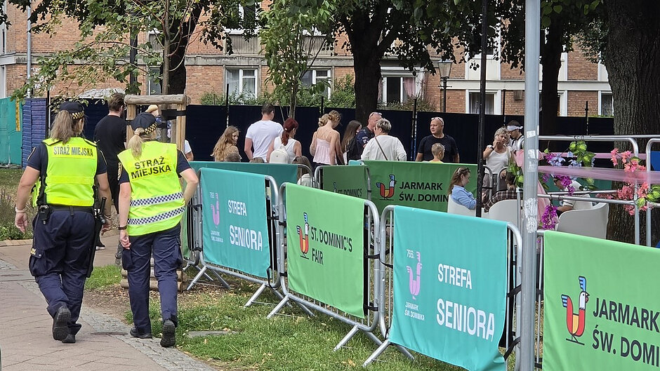 Na zdjęciu widocznych jest dwoje funkcjonariuszy Straży Miejskiej patrolujących teren wydarzenia plenerowego. Funkcjonariusze poruszają się wzdłuż ogrodzenia zabezpieczonego barierkami. Na banerach umieszczonych na barierkach znajdują się napisy „Jarmark Św. Dominika”, St. Dominic's Fair” oraz „Strefa Seniora”. W tle znajdują się uczestnicy wydarzenia korzystający ze strefy wypoczynkowej z ustawionymi stolikami i krzesłami. Otoczenie stanowią drzewa oraz zabudowa miejska, co wskazuje na teren rekreacyjny zorganizowany na potrzeby jarmarku. Zdjęcie dokumentuje działania patrolowe oraz nadzór nad bezpieczeństwem uczestników wydarzenia.