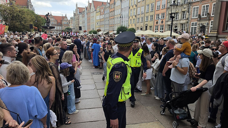 Fotografia przedstawia tłum zgromadzony po obu stronach wyznaczonego ciągu komunikacyjnego na Długim Targu w Gdańsku. W centralnej części zdjęcia widoczni są funkcjonariusze Straży Miejskiej pełniący służbę porządkową podczas wydarzenia publicznego. Uczestnicy obserwują przemarsz lub występ odbywający się w pobliżu Fontanny Neptuna, która jest widoczna w tle. Na zdjęciu znajdują się liczne zabytkowe kamienice charakterystyczne dla historycznego centrum Gdańska. Wśród zgromadzonych osób widoczne są także rodziny z dziećmi oraz osoby korzystające z wózków dziecięcych.