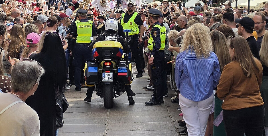 Na zdjęciu przedstawiono działania służb podczas wydarzenia plenerowego na Długim Targu w Gdańsku. W centralnej części kadru znajduje się motocykl ratownictwa medycznego oznakowany napisem „Ratownik Medyczny – Ambulans”. Po obu stronach wyznaczonego przejścia stoją uczestnicy wydarzenia, tworząc korytarz umożliwiający przejazd pojazdu ratunkowego. Wokół motocykla widoczni są funkcjonariusze Straży Miejskiej w kamizelkach odblaskowych, wspierający utrzymanie bezpieczeństwa i drożności ciągów komunikacyjnych. Zdjęcie dokumentuje współpracę służb odpowiedzialnych za bezpieczeństwo uczestników imprezy masowej.