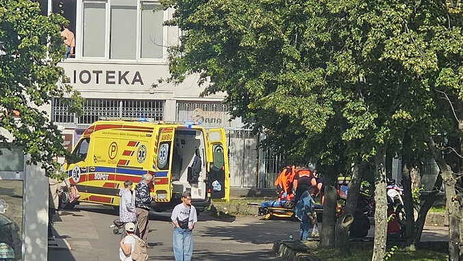 Na zdjęciu widoczny jest ambulans ratownictwa medycznego zaparkowany przy ulicy w rejonie budynku, na którego elewacji znajduje się duży napis „BIBLIOTEKA”. Pojazd ratunkowy jest oznakowany w kolorze żółtym z elementami czerwonymi i niebieskimi, a jego sygnały świetlne są włączone.
W pobliżu ambulansu znajdują się ratownicy medyczni ubrani w odzież o wysokiej widoczności koloru pomarańczowego. Na chodniku obok pojazdu widoczne są nosze przygotowane do transportu osoby wymagającej pomocy medycznej. W tle, częściowo przesłonięte przez drzewa, znajdują się dodatkowe pojazdy służb ratunkowych.
Na pierwszym planie widoczni są piesi poruszający się chodnikiem i przejściem dla pieszych. Miejsce zdarzenia zlokalizowane jest w obszarze miejskim z licznym zadrzewieniem oraz zabudową usługową. Zdjęcie dokumentuje prowadzenie działań ratowniczych przez zespół ratownictwa medycznego w przestrzeni publicznej.