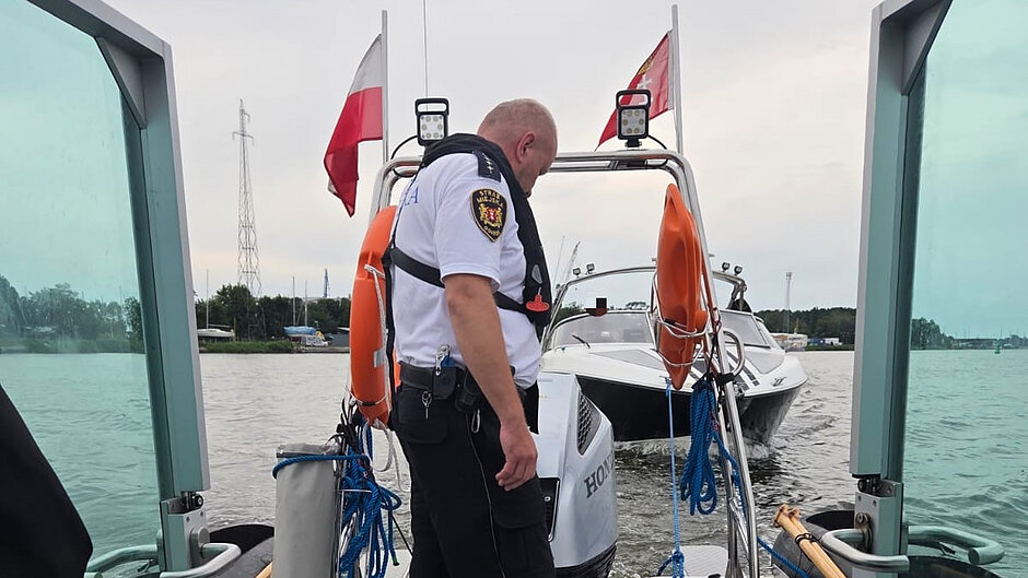 Na zdjęciu widoczna jest jednostka patrolowa znajdująca się na akwenie wodnym. Na rufie łodzi stoi umundurowana osoba wyposażona w kamizelkę ratunkową. Na burcie widoczne są koła ratunkowe, liny cumownicze oraz silnik zaburtowy marki Honda.
Przed jednostką patrolową znajduje się biała motorówka, do której wykonywane jest podejście manewrowe. Na obu jednostkach widoczne są flagi. W tle znajduje się linia brzegowa z drzewami, maszty, infrastruktura portowa oraz słup wysokiego napięcia. Warunki pogodowe są spokojne, niebo jest zachmurzone, a powierzchnia wody lekko pofalowana.