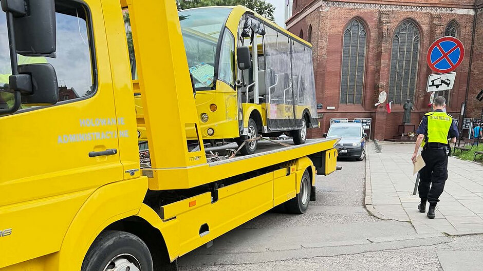 Na zdjęciu widoczny jest żółty pojazd do holowania administracyjnego z załadowanym na platformę niewielkim żółtym pojazdem przypominającym autobus turystyczny. Sytuacja ma miejsce przy zabudowie sakralnej z czerwonej cegły. Po prawej stronie znajduje się osoba w kamizelce odblaskowej z napisem „Straż Miejska”, idąca chodnikiem. Widoczny jest również znak drogowy B-36 „zakaz zatrzymywania się” wraz z tabliczką informującą o możliwości odholowania pojazdu. Na drzwiach holownika znajduje się napis „Holowanie administracyjne”.