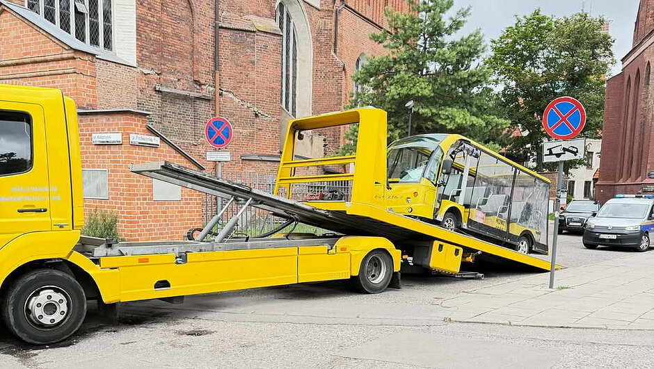 Zdjęcie przedstawia moment załadunku żółtego pojazdu przypominającego autobus turystyczny na lawetę przeznaczoną do holowania administracyjnego. Platforma lawety jest opuszczona i ustawiona pod kątem, co wskazuje na trwającą czynność załadunku lub rozładunku. W tle znajdują się ceglane budynki, w tym kościół, a także zaparkowane pojazdy. W pobliżu widoczny jest znak B-36 „zakaz zatrzymywania się” z tabliczką wskazującą odholowanie pojazdu na koszt właściciela. Całość dokumentuje czynności związane z usunięciem pojazdu z miejsca objętego zakazem zatrzymywania się.