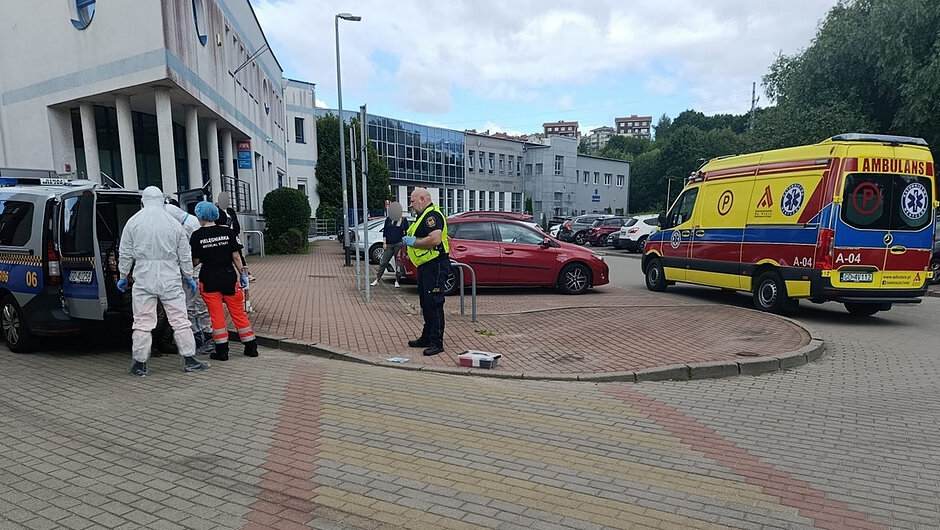 Na zdjęciu widoczny jest teren przed budynkiem użyteczności publicznej. W centralnej części kadru znajduje się utwardzony plac oraz miejsca parkingowe z zaparkowanymi pojazdami. Po lewej stronie widoczny jest samochód służbowy z otwartymi tylnymi drzwiami, przy którym znajdują się osoby uczestniczące w działaniach interwencyjnych. Jedna z osób ubrana jest w biały kombinezon ochronny, a druga w odzież medyczną.
Po prawej stronie zdjęcia zaparkowany jest ambulans ratunkowy w żółto-czerwonym oznakowaniu. W środkowej części kadru znajduje się osoba w kamizelce ostrzegawczej, prowadząca lub koordynująca działania na miejscu zdarzenia. W pobliżu widoczny jest również niewielki pojemnik lub walizka sprzętowa ustawiona na nawierzchni.
W tle znajdują się budynki, zaparkowane samochody osobowe oraz drzewa. Fotografia dokumentuje prowadzenie czynności służbowych przez służby ratownicze i osoby wyposażone w środki ochrony indywidualnej na terenie przed wejściem do budynku.