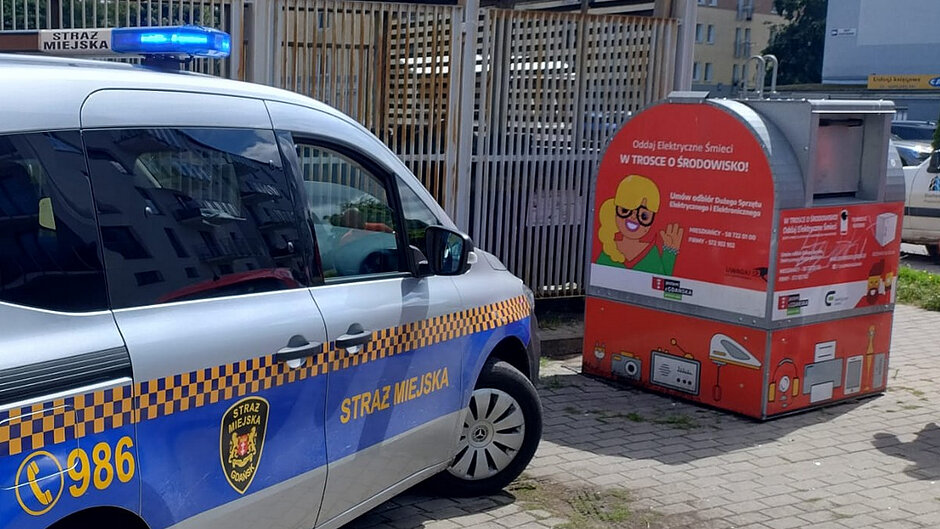 Na zdjęciu widoczny jest oznakowany pojazd Straży Miejskiej zaparkowany obok pojemnika przeznaczonego do zbiórki elektroodpadów. Samochód wyposażony jest w sygnały świetlne, a na drzwiach znajduje się herb Straży Miejskiej oraz numer alarmowy 986. Obok pojazdu ustawiony jest czerwony pojemnik do selektywnej zbiórki zużytego sprzętu elektrycznego i elektronicznego. Na pojemniku widoczny jest napis: „Oddaj elektryczne śmieci w trosce o środowisko!” oraz grafiki przedstawiające przykładowe elektroodpady, takie jak radio, lampka, monitor czy głośnik.
W tle znajduje się zabudowa mieszkaniowa oraz ogrodzona wiata. Zdjęcie zostało wykonane w ciągu dnia, przy dobrej widoczności.
