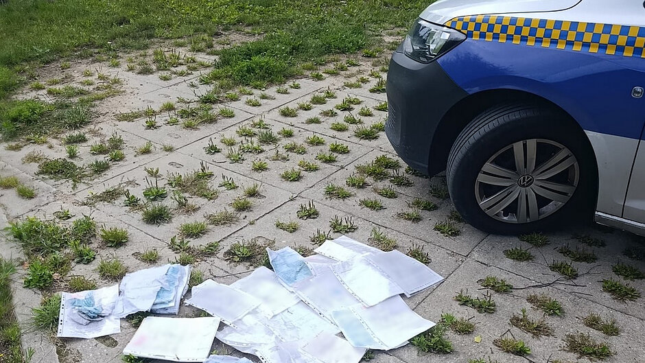 Na zdjęciu widoczny jest fragment utwardzonego terenu z płyt ażurowych porośniętych miejscowo trawą i roślinnością. Po prawej stronie kadru znajduje się zaparkowany samochód służbowy z niebiesko-żółtym oznakowaniem, widoczna jest jego przednia część wraz z kołem. Na pierwszym planie, na nawierzchni obok pojazdu, leżą rozrzucone dokumenty umieszczone w przezroczystych koszulkach foliowych. Część dokumentów jest pognieciona i zabrudzona. Z uwagi na jakość zdjęcia treść dokumentów nie jest czytelna, jednak widoczne są liczne kartki papieru porozrzucane na ziemi. W tle znajduje się teren zielony przylegający do placu. Fotografia dokumentuje pozostawienie lub rozrzucenie dokumentacji papierowej na terenie zewnętrznym w bezpośrednim sąsiedztwie pojazdu służbowego.