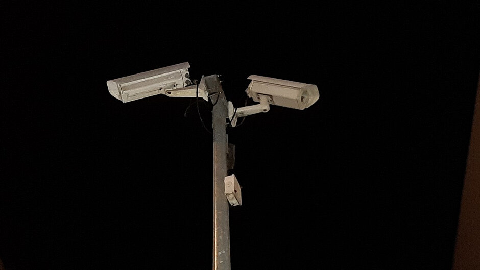 Na zdjęciu widoczny jest system monitoringu wizyjnego zamontowany na metalowym słupie. Do górnej części słupa przymocowane są dwie kamery monitoringu umieszczone po przeciwnych stronach, skierowane w różne kierunki, co pozwala na obserwację większego obszaru. Obudowy kamer mają jasny, kremowo-biały kolor i są osadzone na regulowanych uchwytach. Poniżej kamer znajduje się niewielka skrzynka lub element instalacji elektrycznej zamocowany do słupa. Widoczne są również przewody prowadzące do urządzeń monitorujących.
Zdjęcie wykonano nocą. Tło stanowi ciemne, niemal całkowicie czarne niebo, dzięki czemu głównym elementem fotografii są oświetlone kamery i słup. Po prawej stronie kadru widoczny jest fragment pionowej konstrukcji lub ściany budynku.