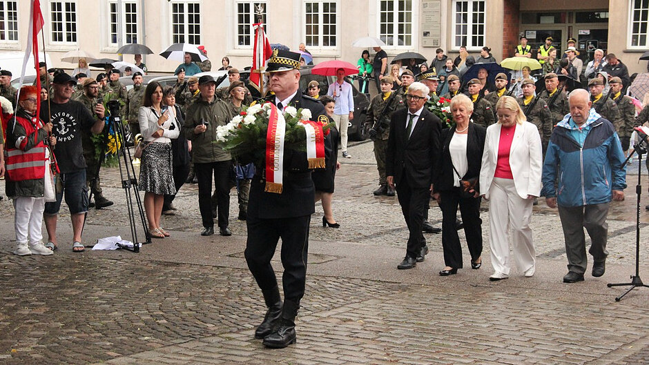 Na zdjęciu widoczny jest przemarsz uczestników uroczystości patriotycznej. Na czele kolumny znajduje się umundurowany funkcjonariusz niosący wieniec ozdobiony biało-czerwonymi wstęgami. Za nim podążają uczestnicy wydarzenia oraz przedstawiciele środowisk kombatanckich, organizacji społecznych i instytucji publicznych. W tle widoczne są pododdziały mundurowe, poczty sztandarowe oraz osoby obserwujące przebieg uroczystości.