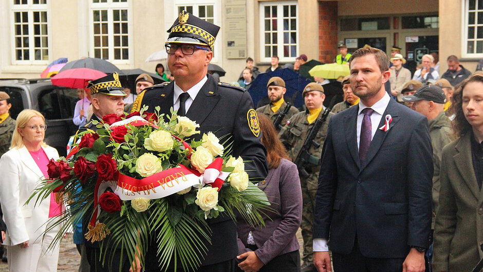 Na zdjęciu widoczny jest moment ceremonii składania wieńca. Umundurowany przedstawiciel służby niesie okazały wieniec wykonany z czerwonych i białych kwiatów, ozdobiony biało-czerwoną szarfą. W tle znajdują się uczestnicy wydarzenia, przedstawiciele służb mundurowych oraz osoby obserwujące uroczystość. Ze względu na opady deszczu część uczestników korzysta z parasoli.