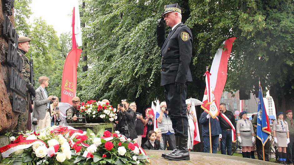 Na zdjęciu widoczny jest fragment uroczystości patriotycznej odbywającej się przy pomniku lub miejscu pamięci. Na pierwszym planie znajduje się umundurowany funkcjonariusz pełniący wartę honorową i oddający salut. Przed pomnikiem złożono liczne wieńce i wiązanki kwiatów w barwach biało-czerwonych. W tle zgromadzeni są uczestnicy uroczystości, poczty sztandarowe oraz przedstawiciele organizacji i służb. Wydarzenie odbywa się na świeżym powietrzu, w otoczeniu drzew, podczas deszczowej pogody.