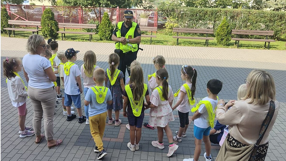 Grupa przedszkolaków wraz z dwiema opiekunkami stoi przed strażnikiem miejskim.