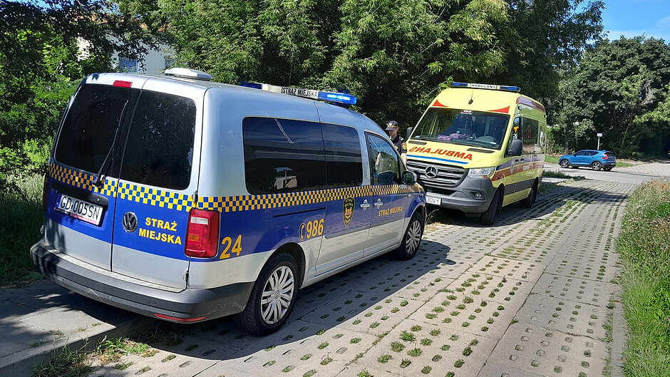 Na zdjęciu widoczne są pojazdy służb ratunkowych zatrzymane na utwardzonej drodze wykonanej z płyt ażurowych, otoczonej zielenią i drzewami. Na pierwszym planie znajduje się oznakowany radiowóz Straży Miejskiej Gdańska marki Volkswagen. Pojazd posiada niebiesko-żółte oznakowanie służbowe, numer boczny 24 oraz oznaczenie G986. Na dachu radiowozu włączone są niebieskie sygnały świetlne.
Przed radiowozem zaparkowana jest karetka pogotowia ratunkowego marki Mercedes-Benz oznakowana napisem „AMBULANS”. Karetka również ma uruchomione sygnały świetlne. Pomiędzy pojazdami widoczna jest osoba uczestnicząca w prowadzonych działaniach.
W tle znajdują się gęste drzewa i krzewy, a po prawej stronie widoczny jest parking z zaparkowanym samochodem osobowym. Warunki pogodowe są dobre, a zdjęcie wykonano w słoneczny dzień.
Fotografia dokumentuje wspólne działania Straży Miejskiej i zespołu ratownictwa medycznego podczas interwencji prowadzonej w terenie miejskim.