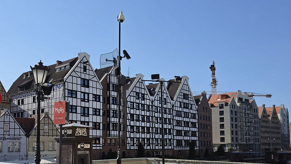 Na zdjęciu przedstawiono fragment zabudowy Śródmieścia Gdańska nad brzegiem Motławy. W centralnej części kadru widoczne są charakterystyczne budynki o szczytowych dachach i elewacjach stylizowanych na historyczną architekturę gdańską. Fasady budynków utrzymane są w biało-czarnej kolorystyce, z wyraźnie zaznaczoną konstrukcją ryglową. W dalszej części widoczne są nowsze budynki mieszkalne oraz żurawie budowlane świadczące o prowadzonych pracach inwestycyjnych.

Na pierwszym planie znajduje się słup wyposażony w elementy monitoringu miejskiego. Na jego szczycie zamontowana jest obrotowa kamera monitoringu, poniżej widoczne są dodatkowe urządzenia obserwacyjne oraz oświetlenie. Obok znajduje się stylizowana latarnia uliczna. Widoczne wyposażenie wskazuje na funkcjonowanie systemu monitoringu służącego obserwacji przestrzeni publicznej.

W dolnej części zdjęcia znajduje się urządzenie techniczne z widocznym napisem „Gdańsk Główny” oraz baner reklamowy z napisem „MeVo”. Zdjęcie wykonano w słoneczny dzień przy bezchmurnym niebie, co zapewnia dobrą widoczność elementów infrastruktury miejskiej i zabudowy znajdującej się w tle.
Fotografia dokumentuje miejską infrastrukturę monitoringu oraz otaczającą ją zabudowę zlokalizowaną w historycznej części Gdańska.