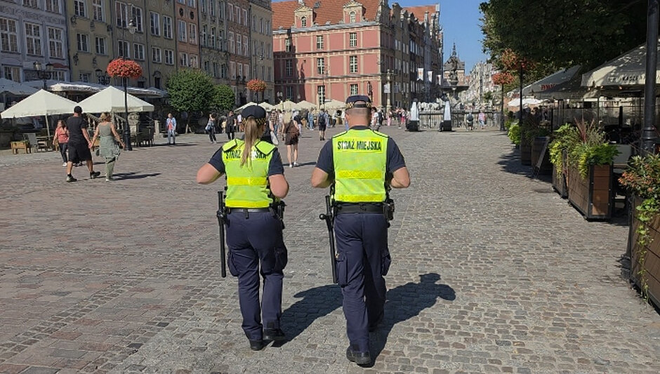 Na zdjęciu widocznych jest dwoje funkcjonariuszy Straży Miejskiej patrolujących rejon Długiego Targu w Gdańsku. Funkcjonariusze ubrani są w granatowe mundury oraz kamizelki ostrzegawcze z napisem „STRAŻ MIEJSKA”. Poruszają się po brukowanej nawierzchni historycznego centrum miasta. W tle znajdują się zabytkowe kamienice charakterystyczne dla Głównego Miasta, ogródki gastronomiczne oraz liczni mieszkańcy i turyści korzystający z przestrzeni publicznej. W centralnej części kadru widoczna jest Fontanna Neptuna. Zdjęcie dokumentuje działania patrolowe prowadzone przez Straż Miejską w rejonie o dużym natężeniu ruchu pieszego.