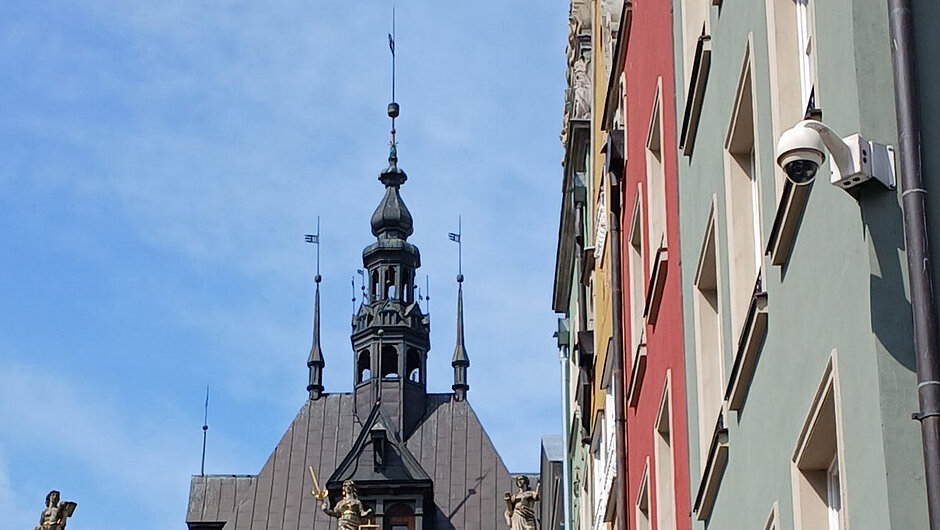 Na zdjęciu przedstawiono kamerę monitoringu miejskiego zamontowaną na elewacji jednej z kamienic w historycznej części Gdańska. Kamera umieszczona jest na wysięgniku przymocowanym do budynku i skierowana w stronę przestrzeni publicznej. W tle widoczna jest wieża zabytkowego ratusza oraz kolorowe fasady sąsiednich kamienic. Zdjęcie wykonano przy dobrej widoczności, co pozwala na wyraźne zobrazowanie elementów infrastruktury monitoringu oraz otaczającej zabudowy miejskiej. Fotografia dokumentuje funkcjonowanie systemu monitoringu wykorzystywanego do obserwacji i poprawy bezpieczeństwa w przestrzeni publicznej.
